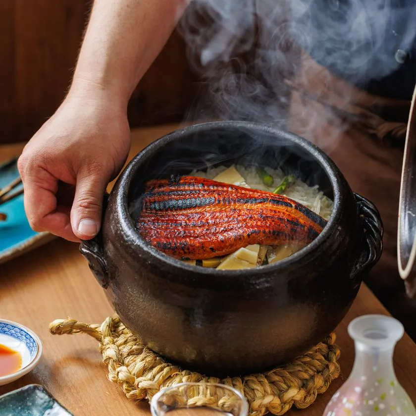 黒い土鍋に炭火焼きの鰻蒲焼きを丸ごと1枚乗せた鰻土鍋飯の提供シーン。湯気が立ち、徳利と小皿が卓上に並ぶ