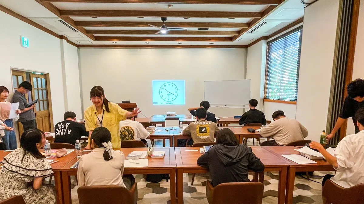 ホテル内研修室でスタッフが時計の絵をスクリーンに映しながら参加者約10名に説明している室内研修の場面