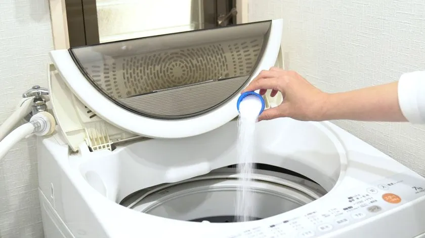 縦型全自動洗濯機の投入口に、付属の計量カップで白色粉末洗剤を注ぎ入れている使用シーン写真。オキシクリーン粉末タイプの実際の使い方を示す