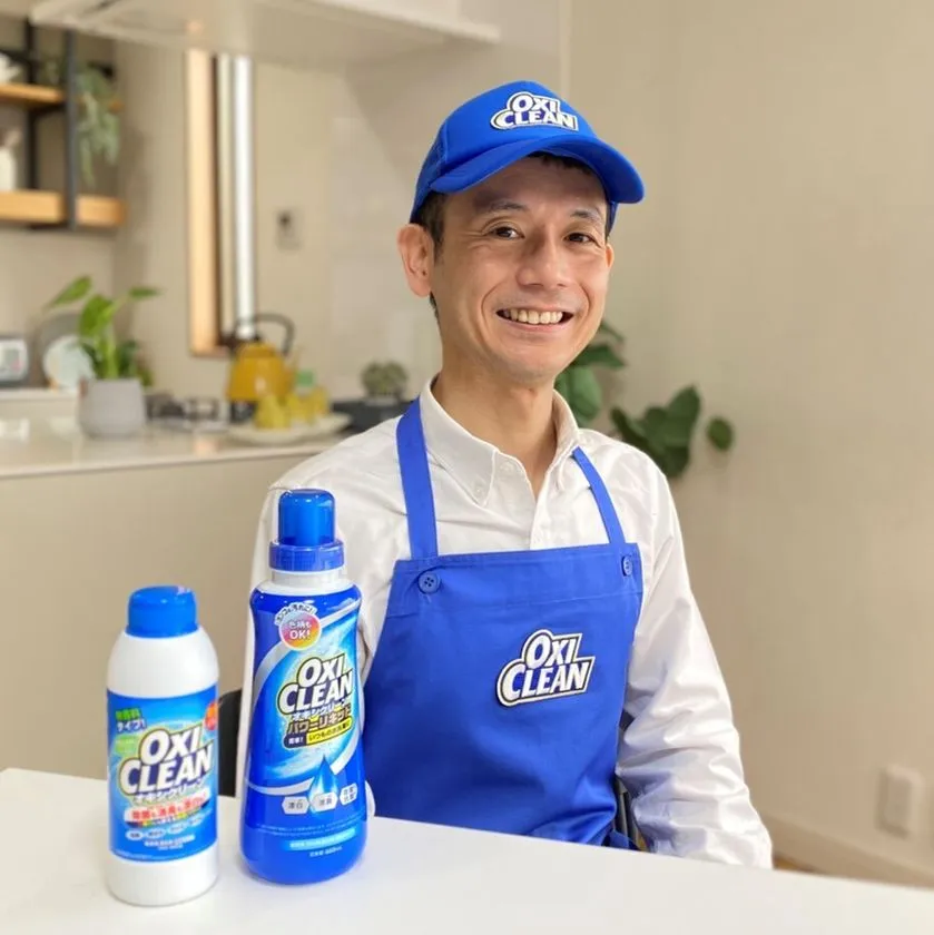 オキシクリーンのロゴ入り青いエプロンとキャップを着用した男性が、粉末タイプと液体タイプのオキシクリーン2製品をテーブルに並べてキッチン背景で笑顔でポーズをとっている広報写真