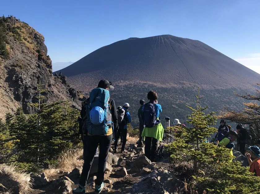 登山者グループが岩場の登山道を浅間山（前掛山）山頂方向へ向かう秋季の登山シーン。バックパックを背負った複数名が列をなして歩く。