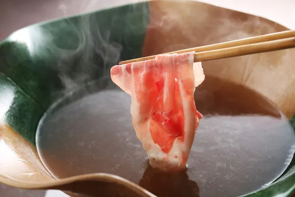 箸でつまみ上げた豚しゃぶ肉の薄切りスライスが湯気を上げる瞬間をクローズアップした料理写真。緑釉の器に入った出汁スープが背景に写る