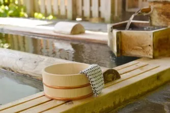 露天風呂の湯船縁に置かれた木製の湯桶と手ぬぐい、竹筒からの湯の流れ、背景に木製の囲い。温泉情景の写真