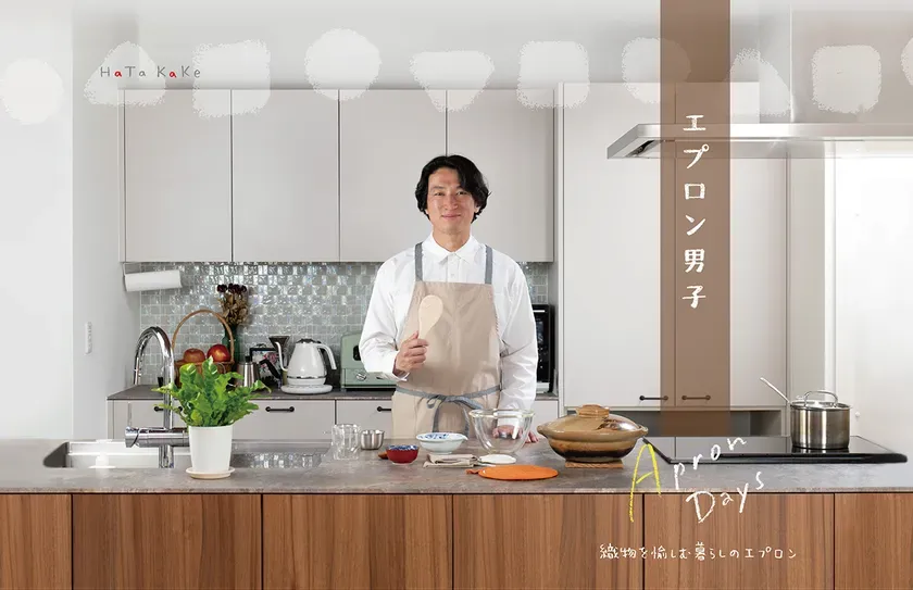 白いキッチンに立つ男性がベージュ系エプロンを着用し、調理道具