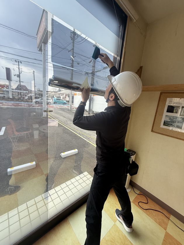 作業員がヘルメットを着用して店舗のガラス窓に遮熱フィルムを施工している実演の様子