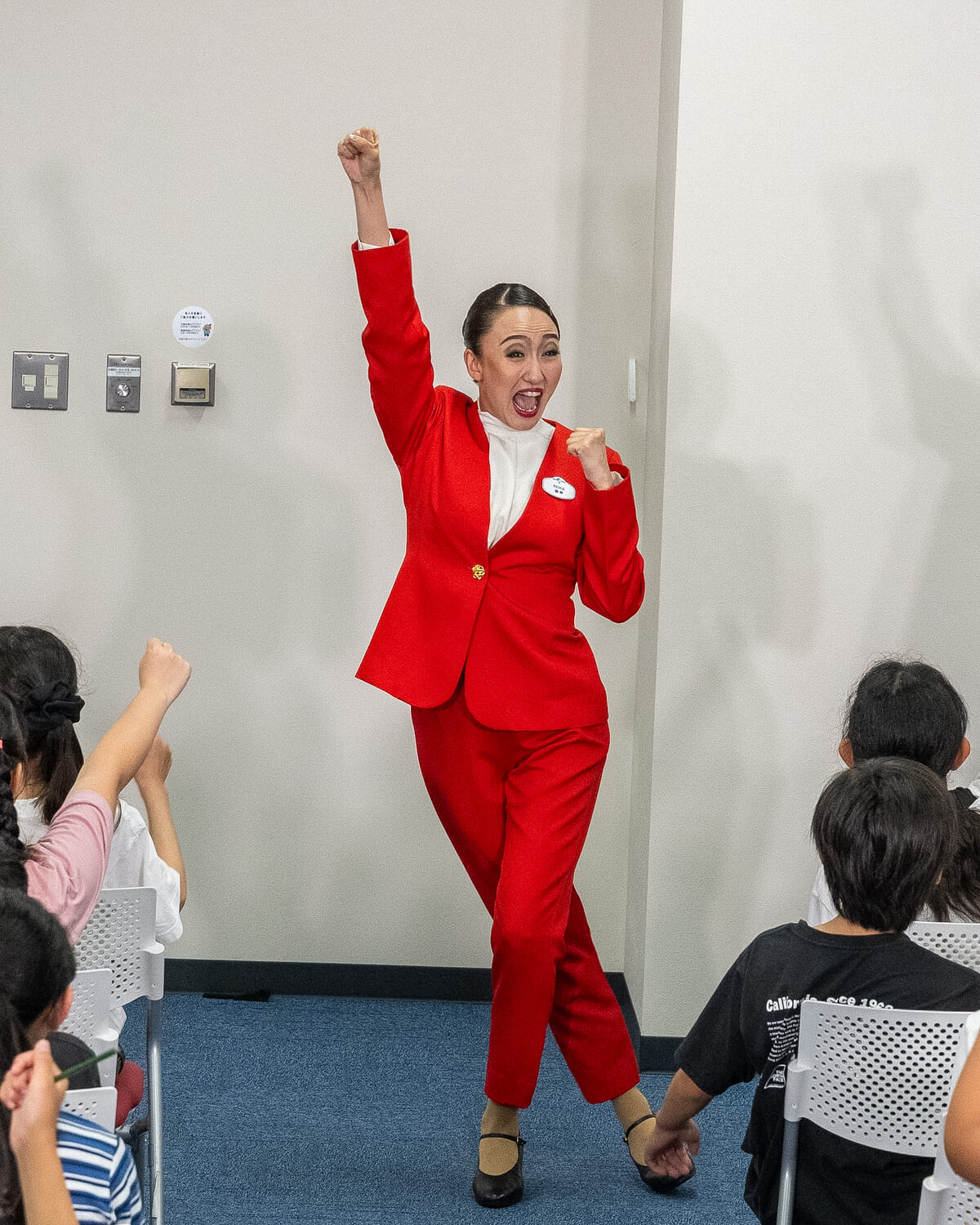 赤いスーツの「綾小路麗華」が片手を高く上げ、子どもたちも手を挙げて応える様子
