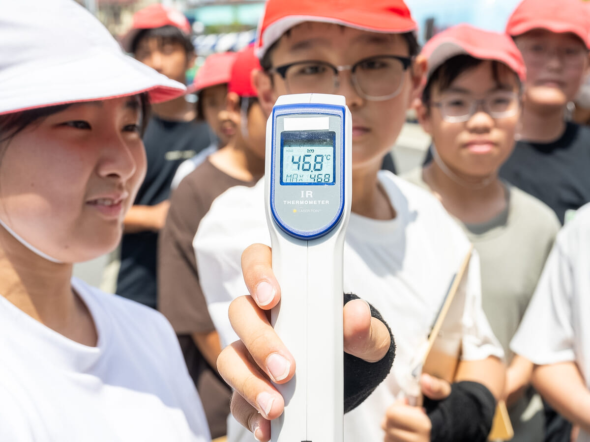 子どもが手に持つ赤外線温度計が日ざしの下で46.8度を示す様子