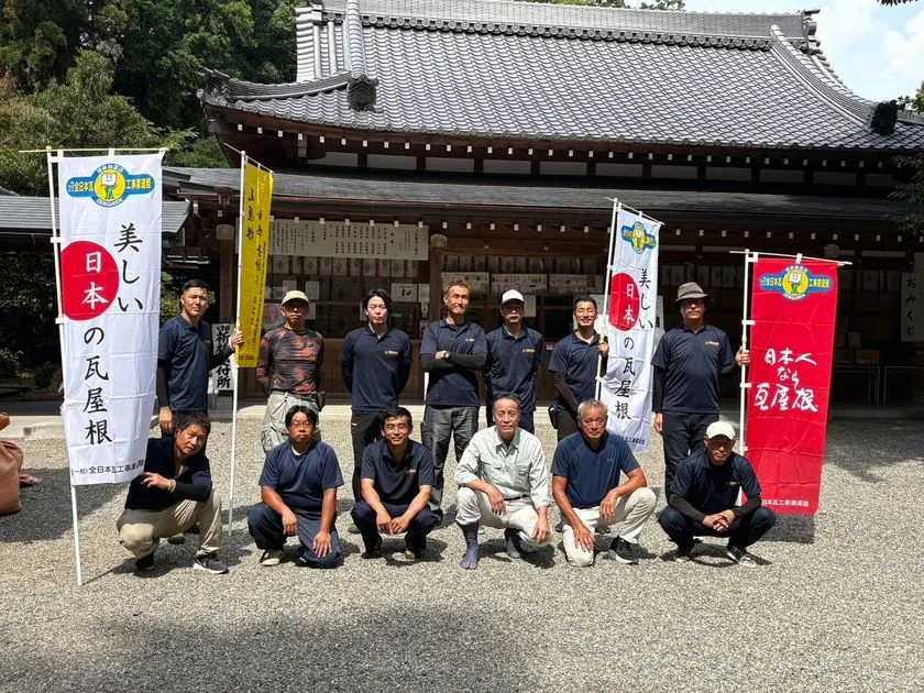 寺院建築の瓦屋根を背景に、のぼり旗を囲んで集合する作業着姿の人々