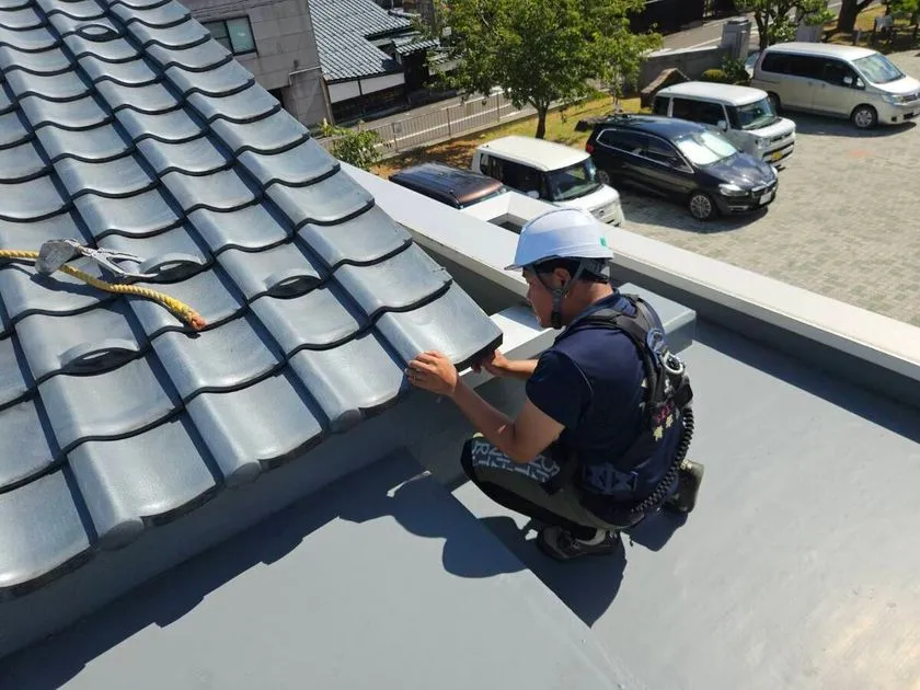 安全帯を装着した職人が、住宅の灰色の瓦屋根の端部を手で点検している