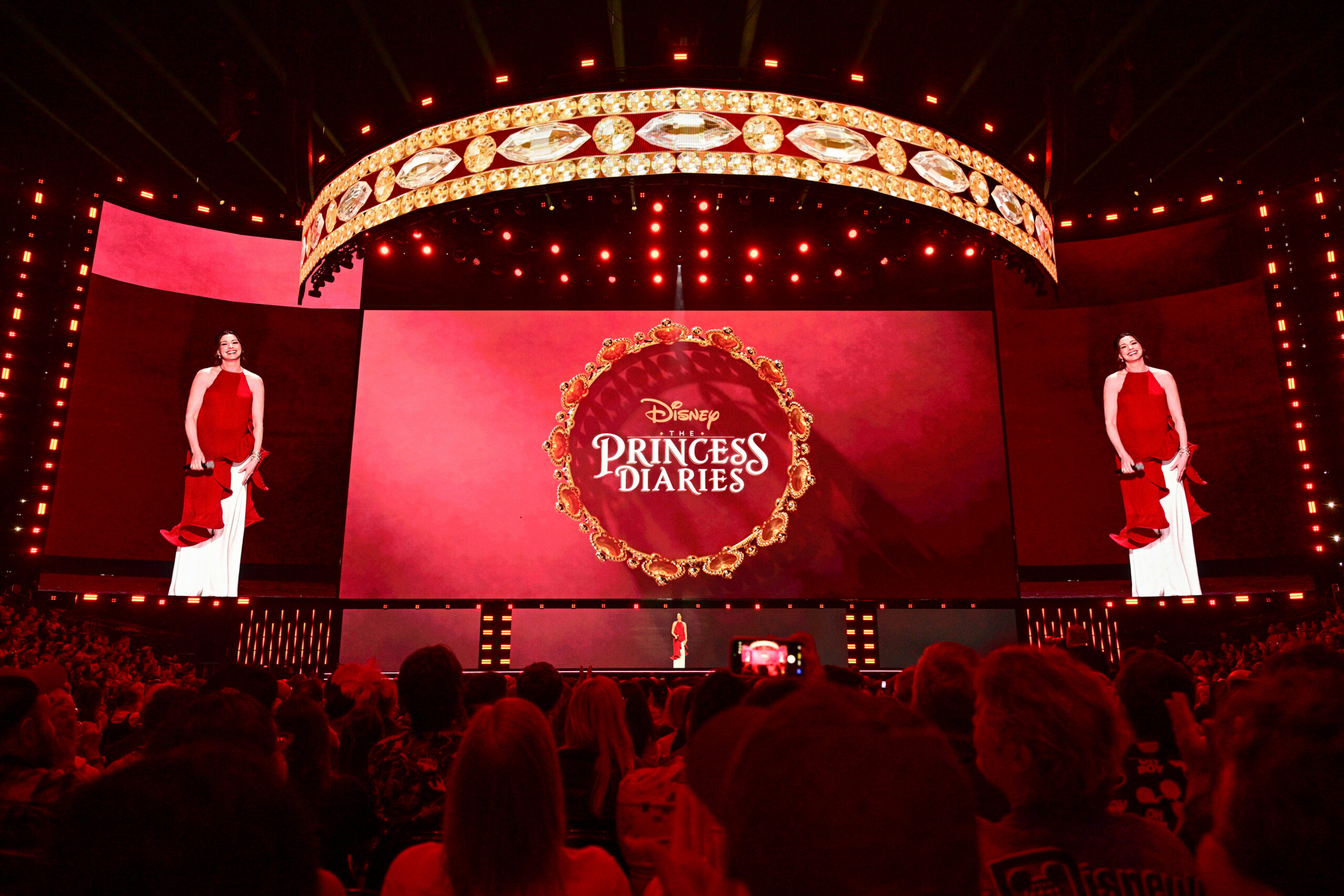 The Princess Diariesのロゴを背にDisney Entertainment Showcaseのステージに立つアン・ハサウェイ