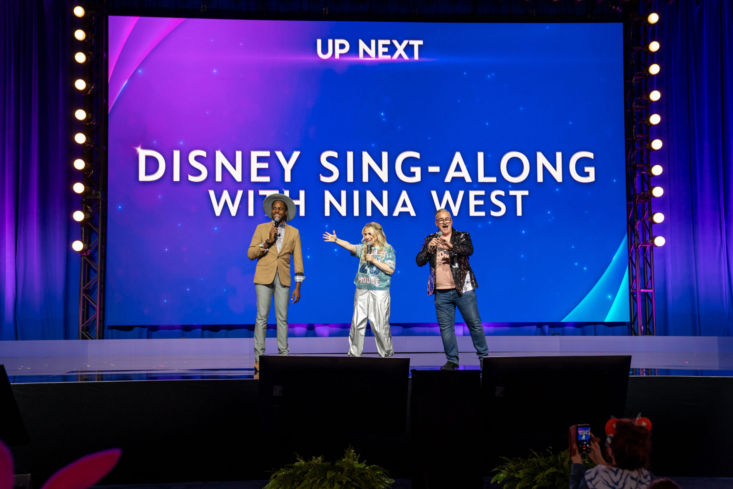 「DISNEY SING-ALONG WITH NINA WEST」の告知が映る大型スクリーンを背に立つアシュリー・エクスタイン、レイヴォン・レディング、ダニー・スタイルズ
