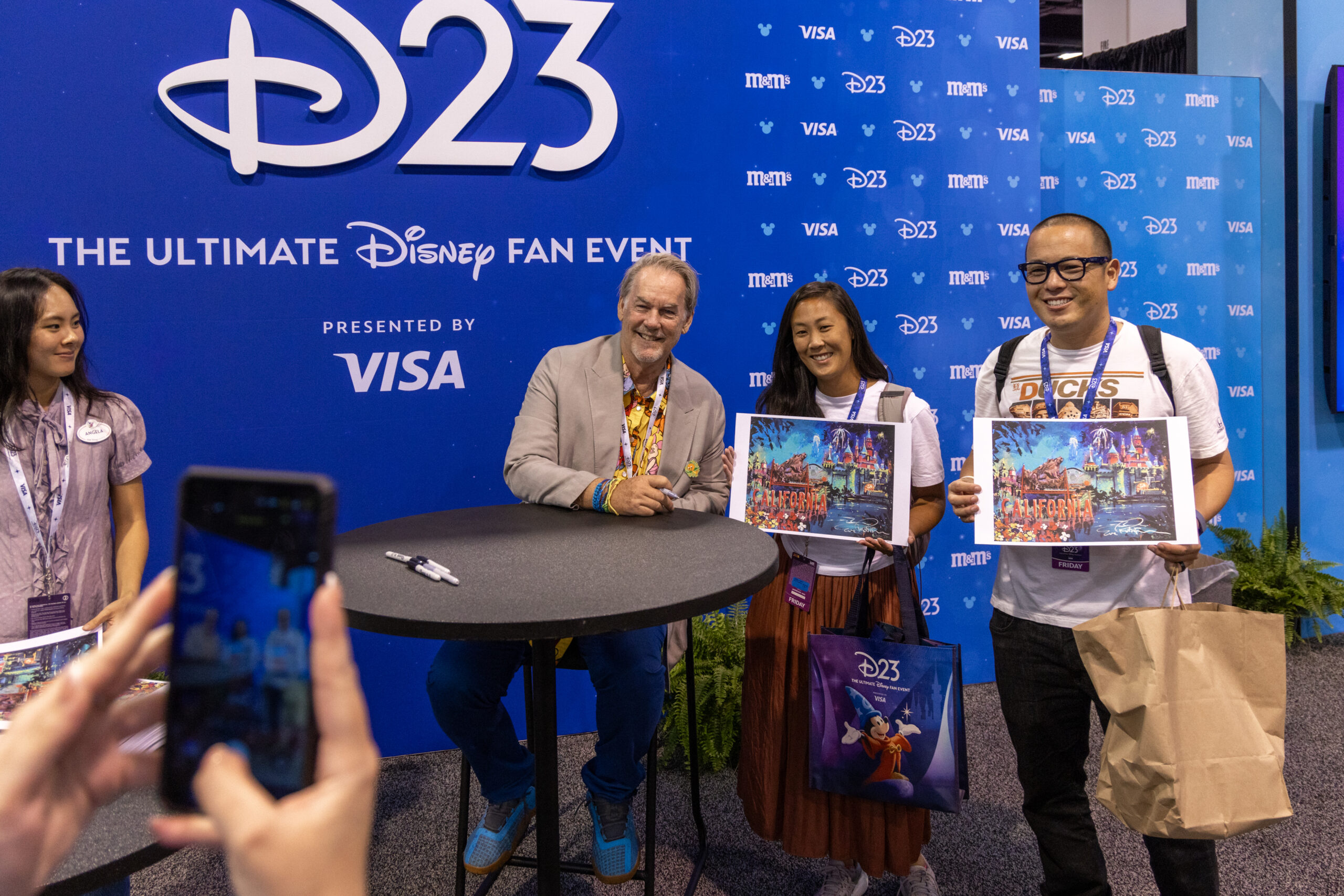 D23のバックドロップの前で、サイン入りの印刷物を持つ3人のファンと並ぶエリック・ロビソン