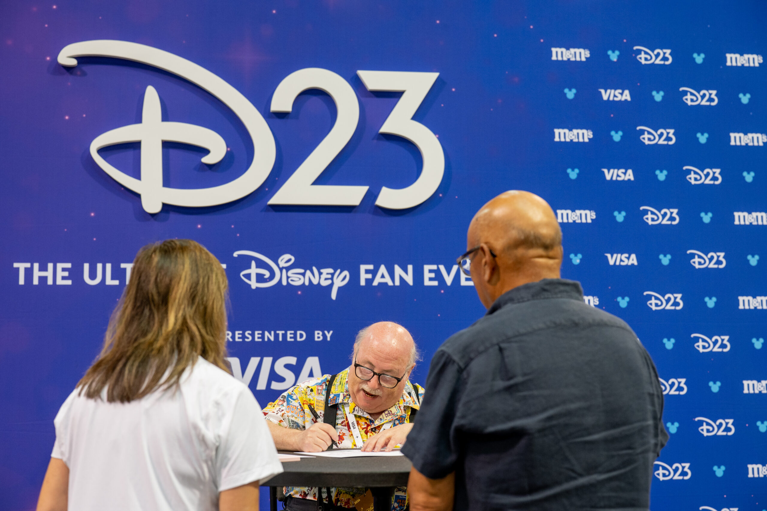 D23の大型ロゴが掲げられた壁面の前で、テーブルに着いてサインを書き入れるエリック・ゴールドバーグ