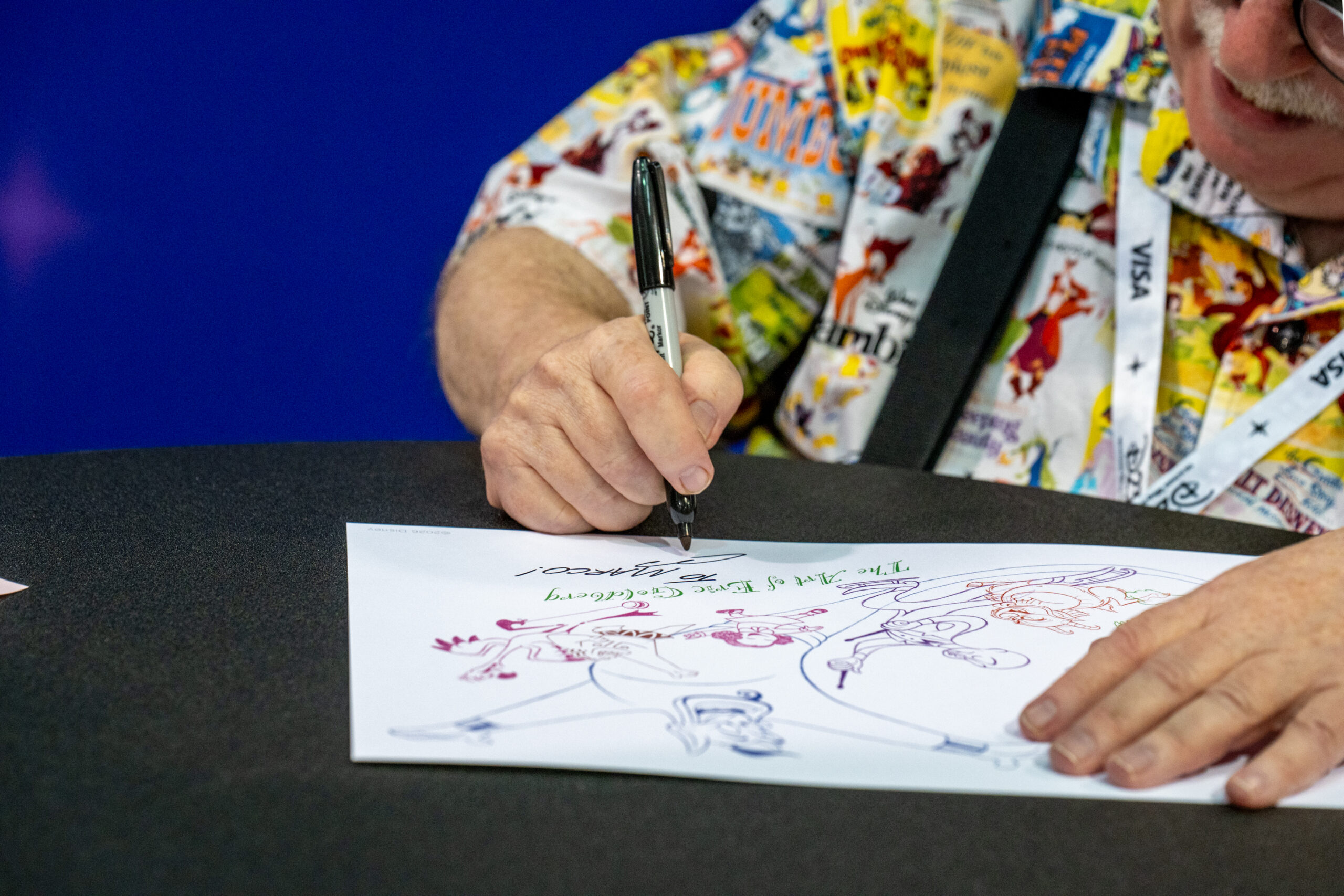 多色の線画キャラクターが描かれたイラストに、ペンでサインを書き入れるエリック・ゴールドバーグの手元