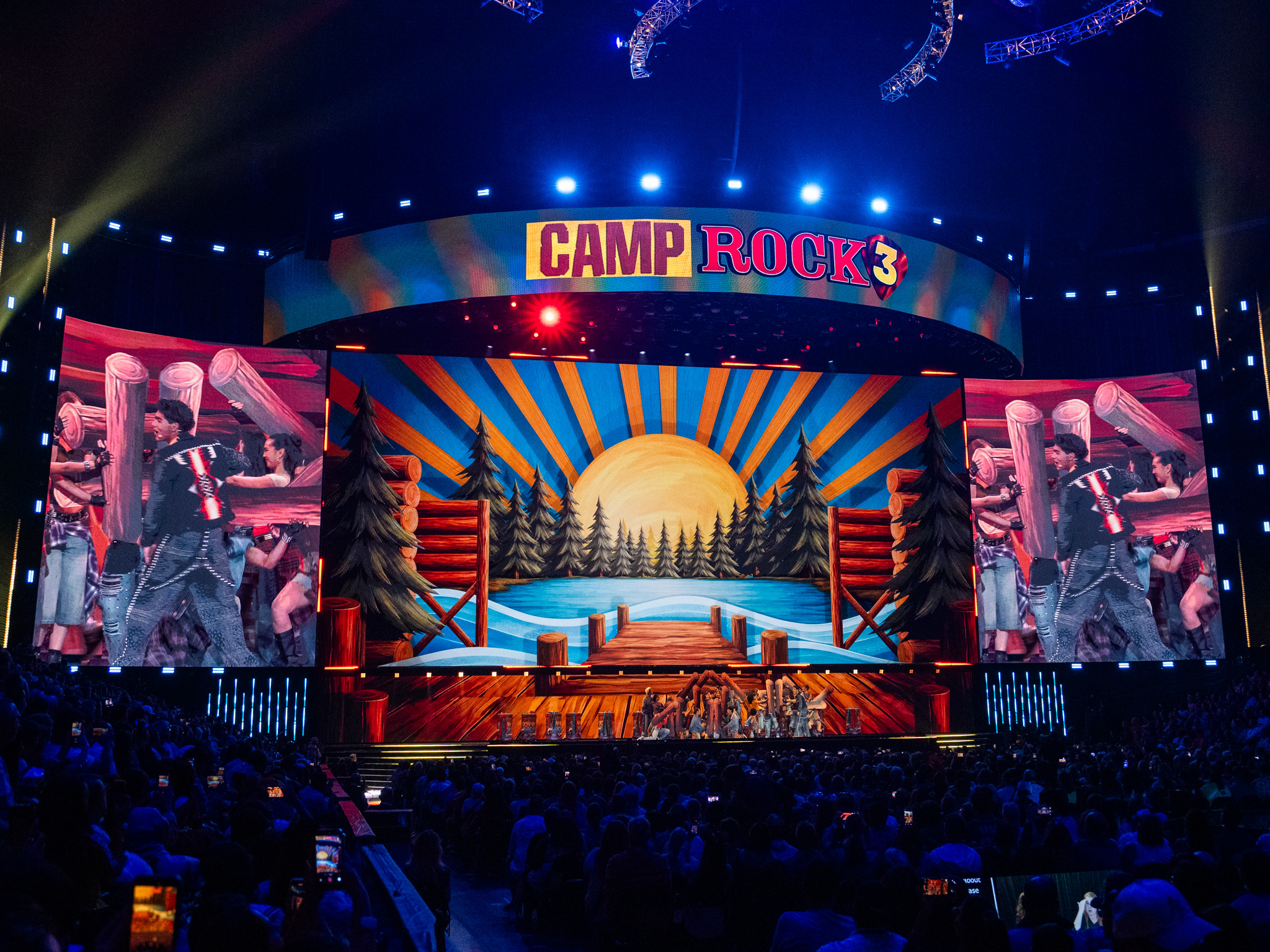 湖と桟橋を描いたキャンプのセットで歌う出演者たちとCAMP ROCK 3のロゴ