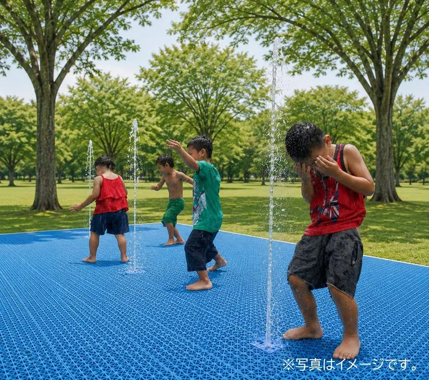 芝生と並木に囲まれた屋外で、子どもたちが青い床面から上がる細い噴水で水遊びをしているイメージ