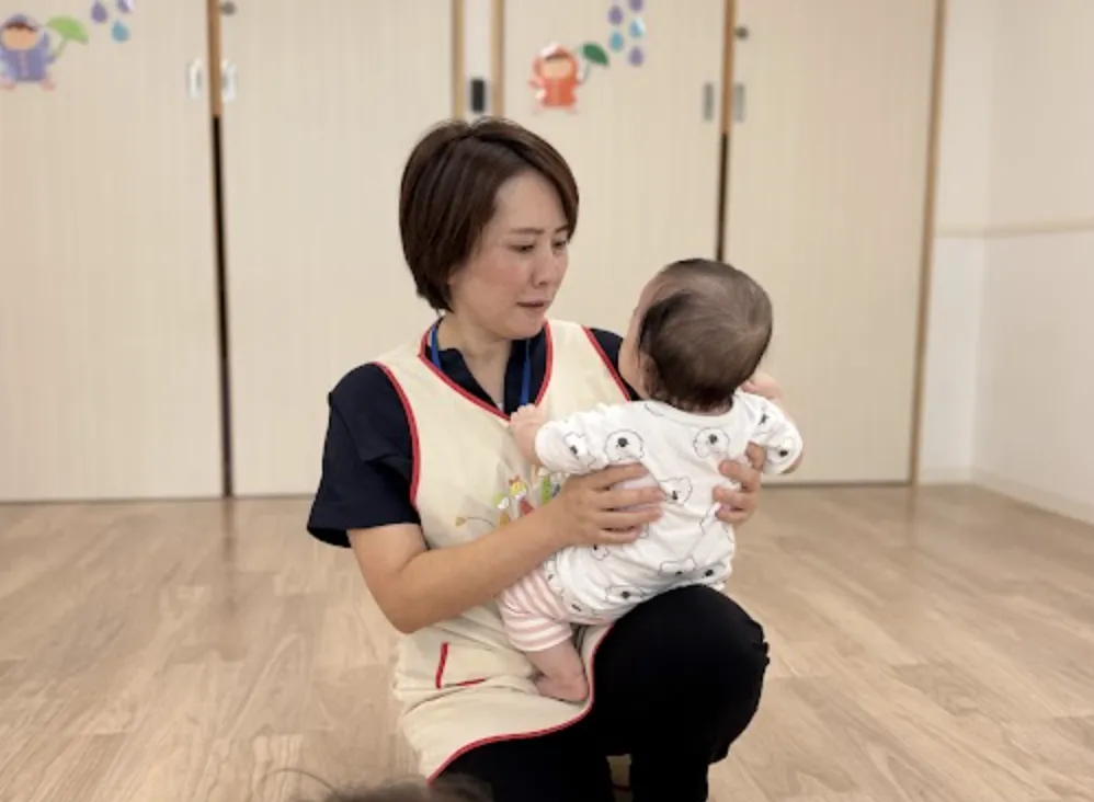 エプロン姿の保育士が赤ちゃんを抱き上げる様子