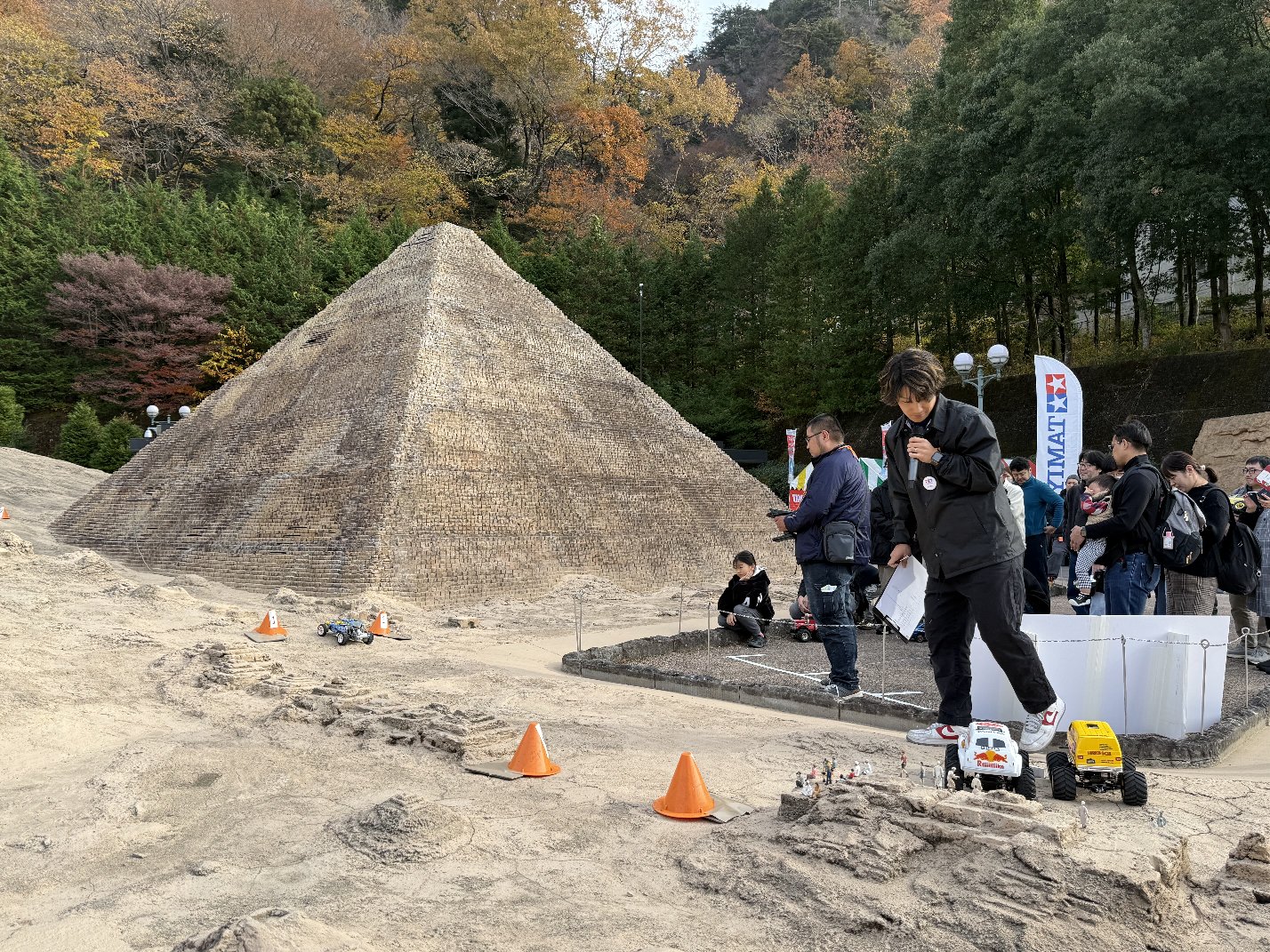 巨大な白いピラミッド模型を背景に砂地のコースでRCカーを走らせる参加者と観客