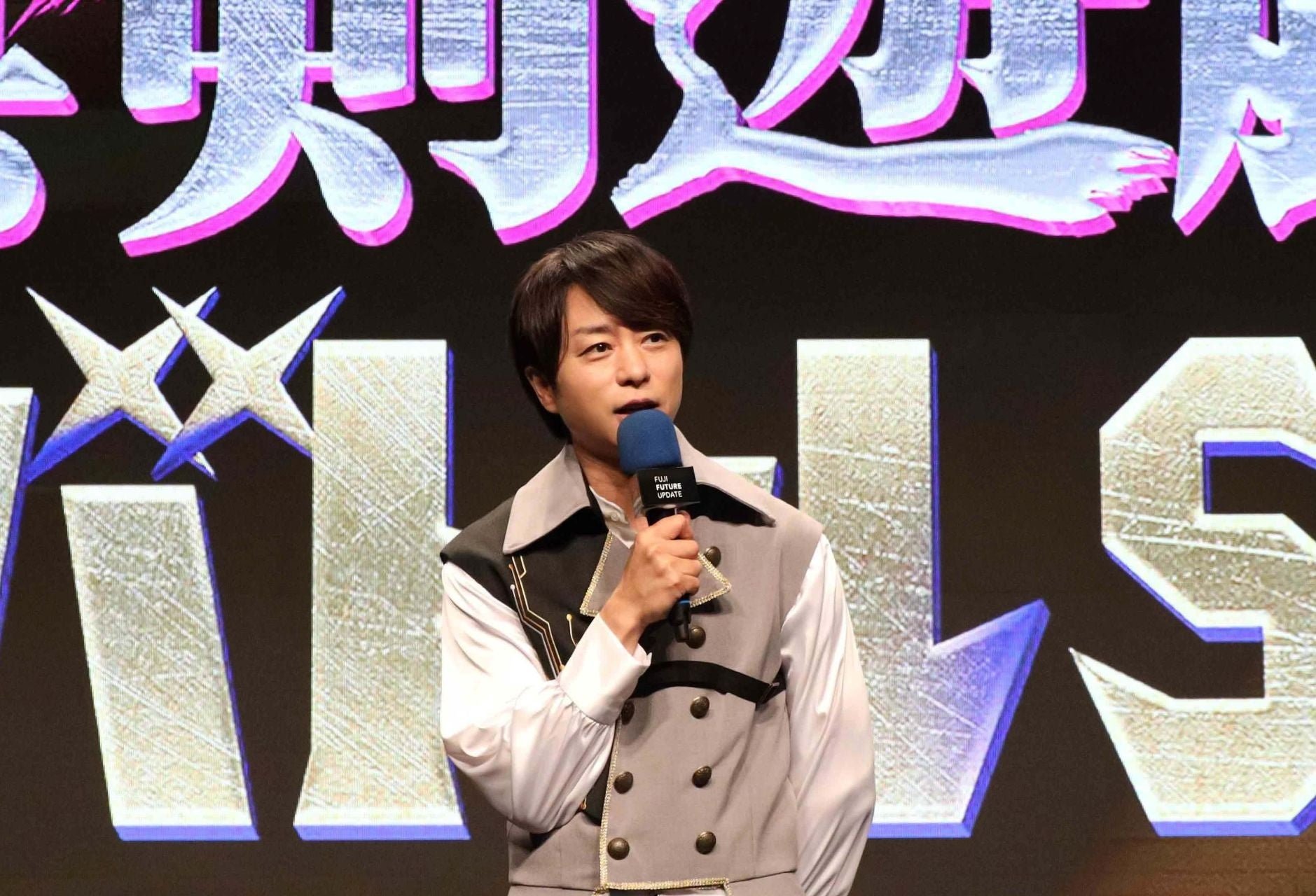 『真剣遊戯！THEバトルSHOW』の告知ビジュアル前でマイクを持つ櫻井翔さん
