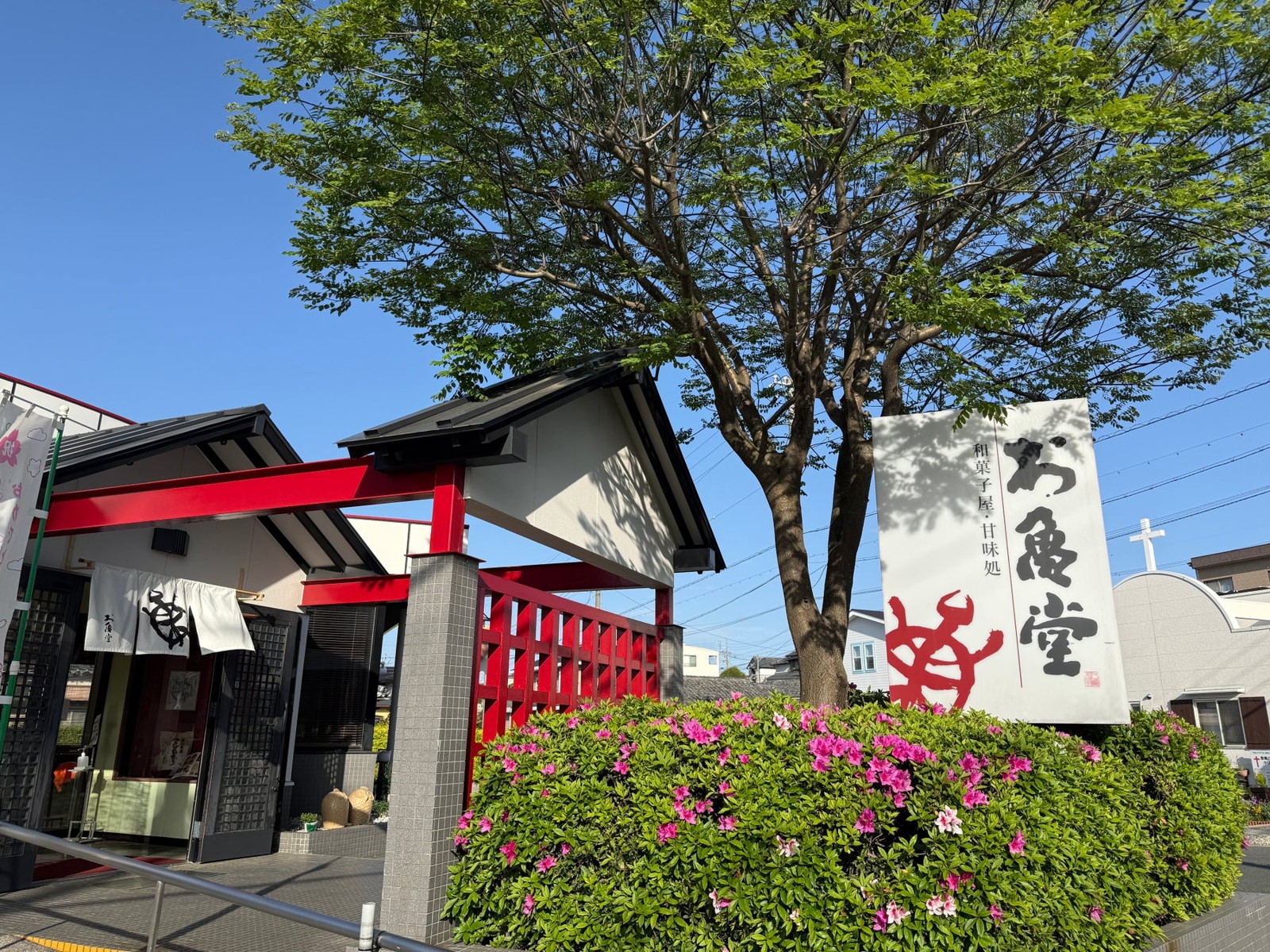 「お亀堂」の看板と赤い門構えのある店舗外観、つつじの花と木々