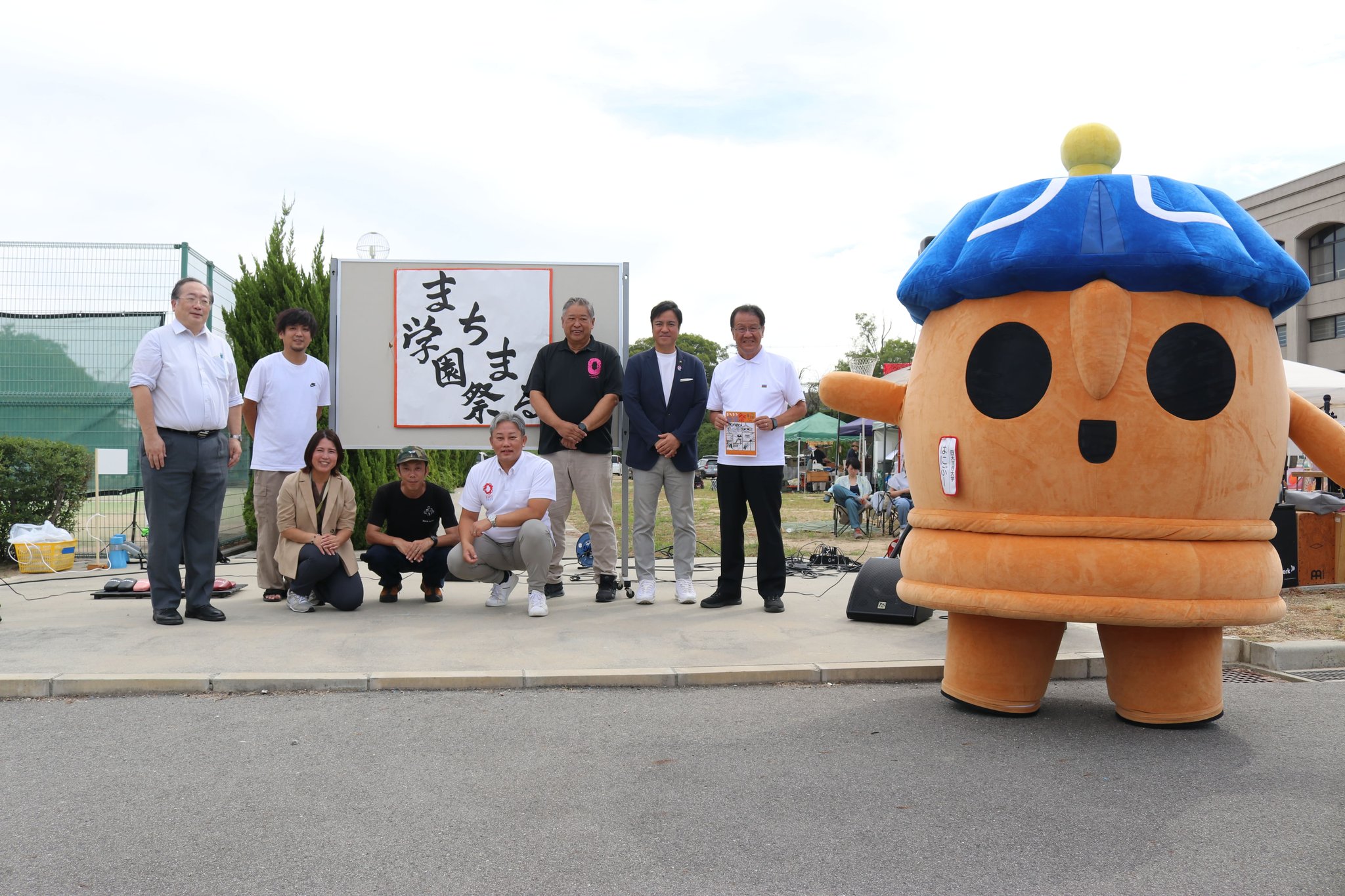 「まちまる学園祭る」看板前に集うマスコットと関係者