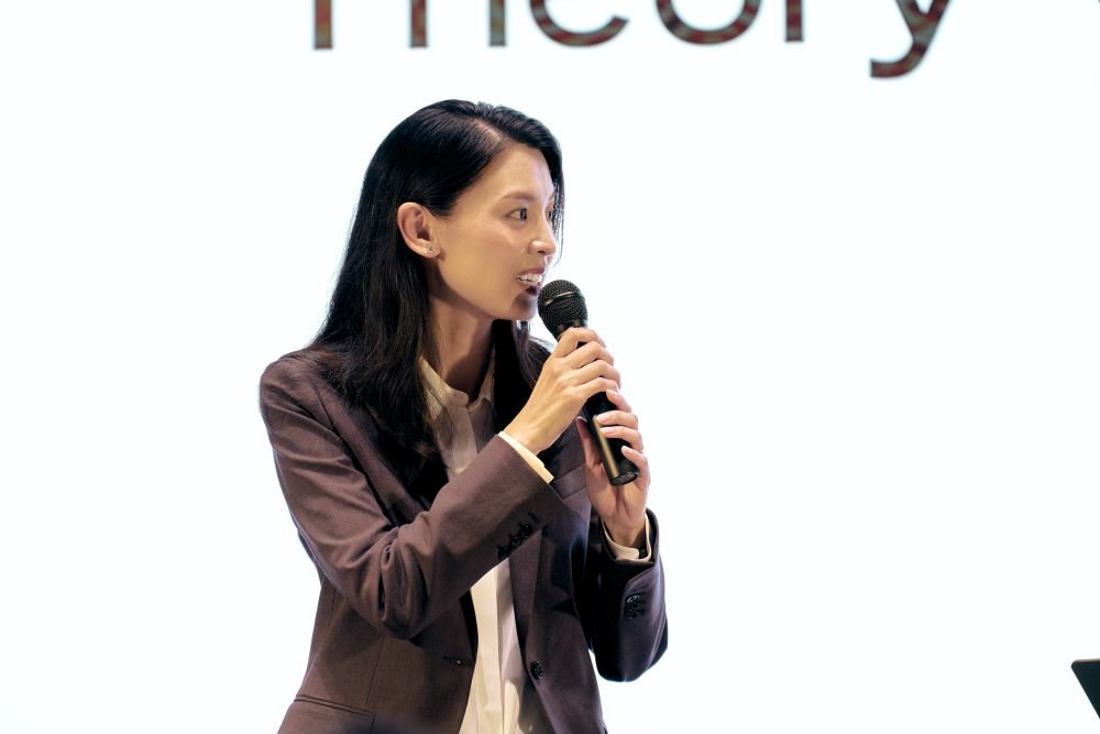 申真衣さんがマイクを持ちTheoryのロゴが映るスクリーン前で話す様子