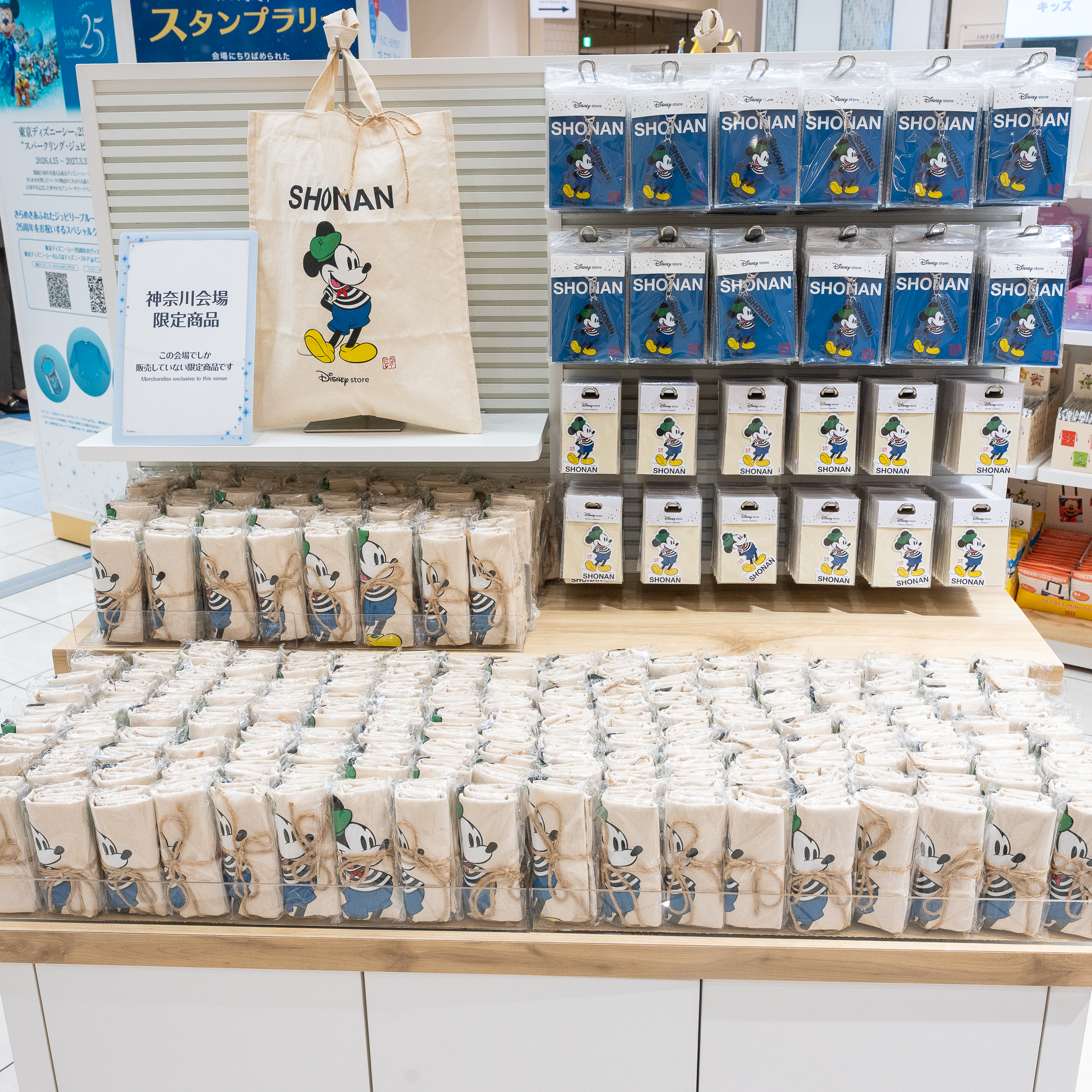 神奈川会場限定商品の案内と、SHONANの文字入りのトートバッグ、キーチェーン、ステッカーが並ぶ売り場