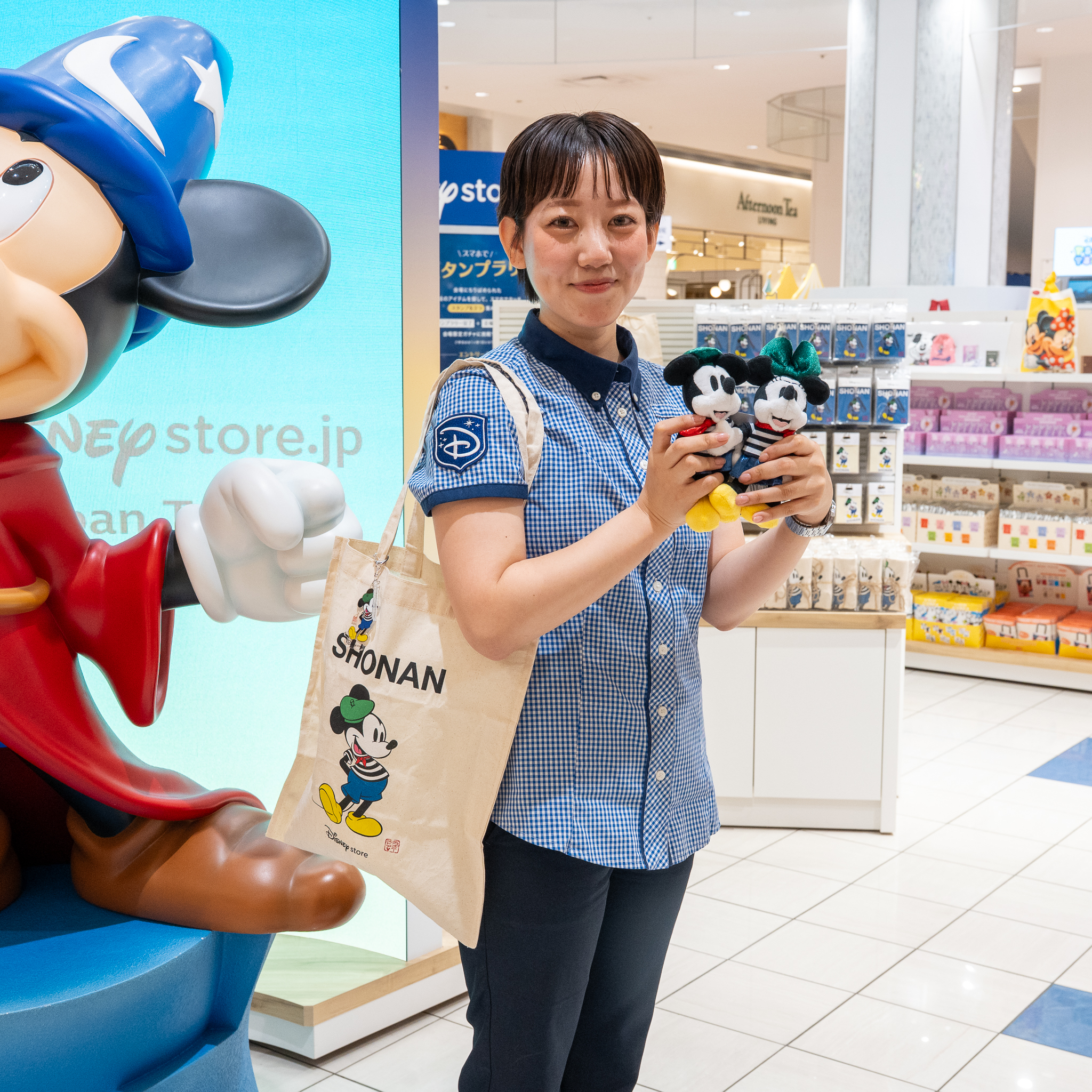 ディズニーストアの会場で、SHONANのトートバッグとミッキーマウスとミニーマウスのぬいぐるみを持つキャストと、魔法使いの弟子に扮したミッキーマウスのスタチュー