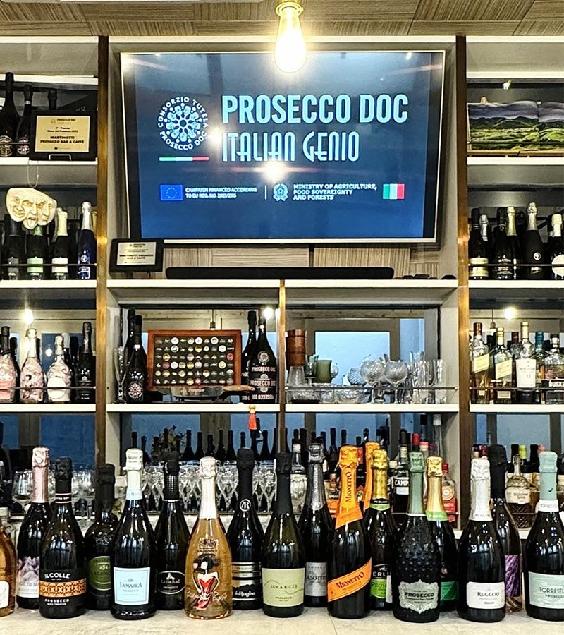 PROSECCO DOCの画面とワインボトルが並ぶ店内