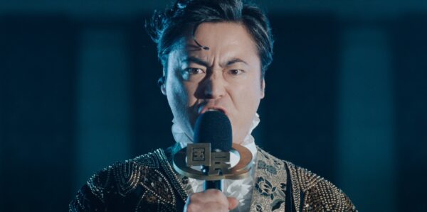 Netflixシリーズ「国民クイズ」の新規場面カット（白いフリル襟と金色の装飾の衣装でマイクを握る場面）