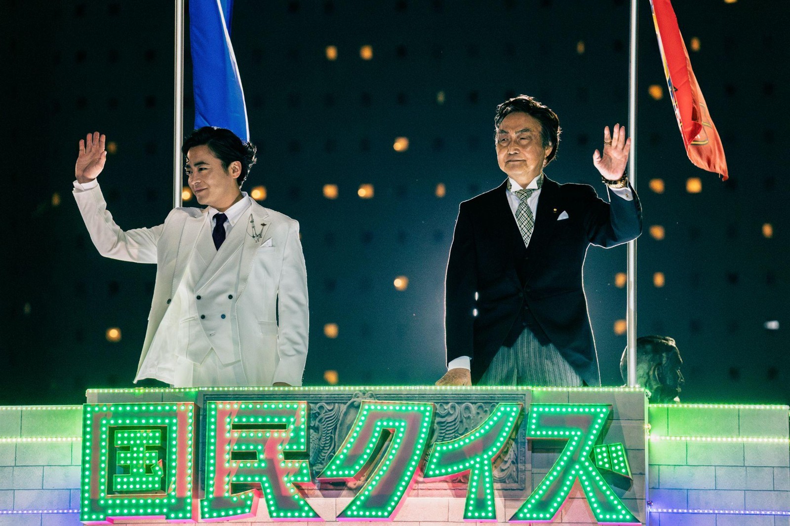Netflixシリーズ「国民クイズ」の新規場面カット（白いタキシード姿のK井K一が総理大臣・獄間沢と並んで手を振る場面）